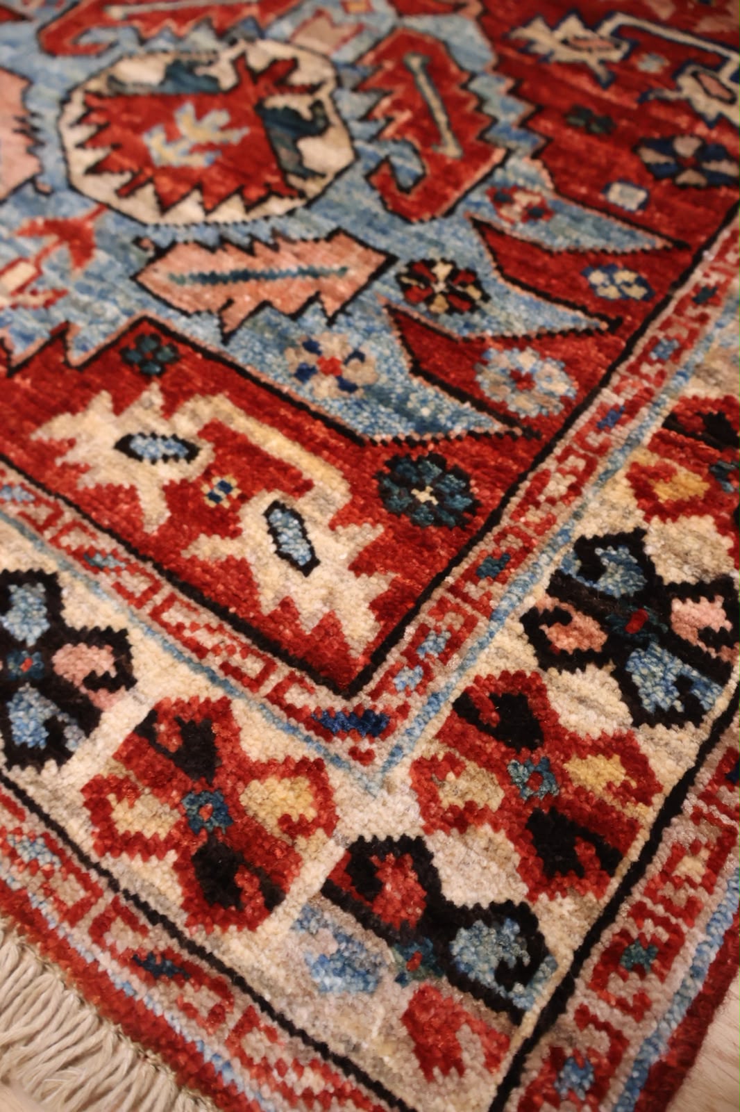 A close-up of a traditional handwoven rug showing its detailed pattern and texture. The rug features a complex geometric design with red, blue, beige, and black motifs, including a prominent cream-colored fringe at the bottom left.