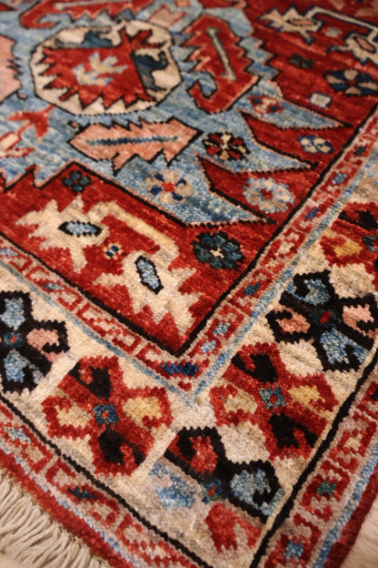 A close-up of a traditional handwoven rug showing its detailed pattern and texture. The rug features a complex geometric design with red, blue, beige, and black motifs, including a prominent cream-colored fringe at the bottom left.