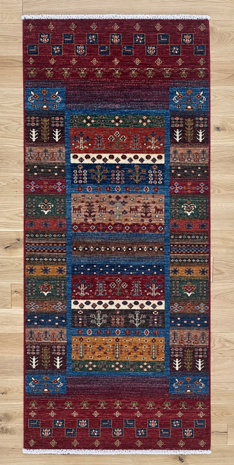 A rectangular patterned area rug with a traditional geometric and tribal design, featuring a deep red border and a central multi-colored panel, shown flat on a light wood floor.