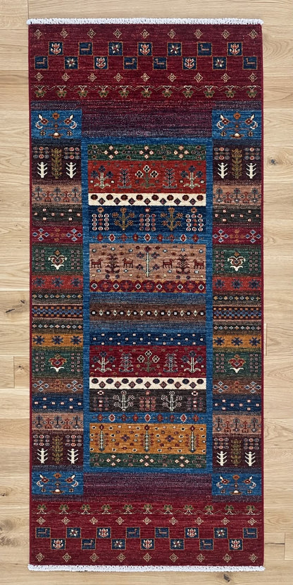 A rectangular patterned area rug with a traditional geometric and tribal design, featuring a deep red border and a central multi-colored panel, shown flat on a light wood floor.