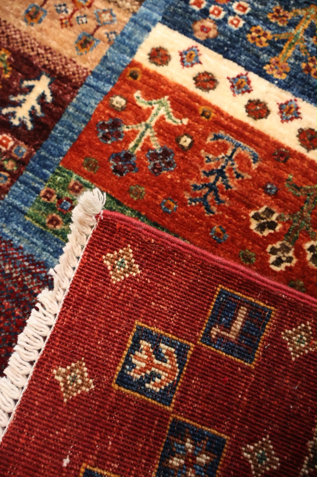 Close-up of a patterned area rug with multiple colors including red, blue, beige, and green, showing detailed woven texture and white fringe.