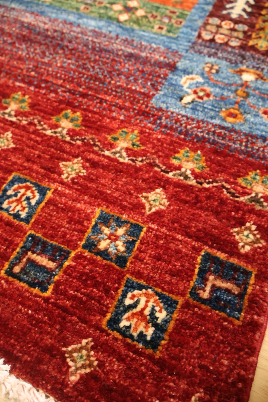 A close-up of a traditional patterned rug with a dominant deep red field, featuring blue squares with cream motifs and a light blue section with floral details.