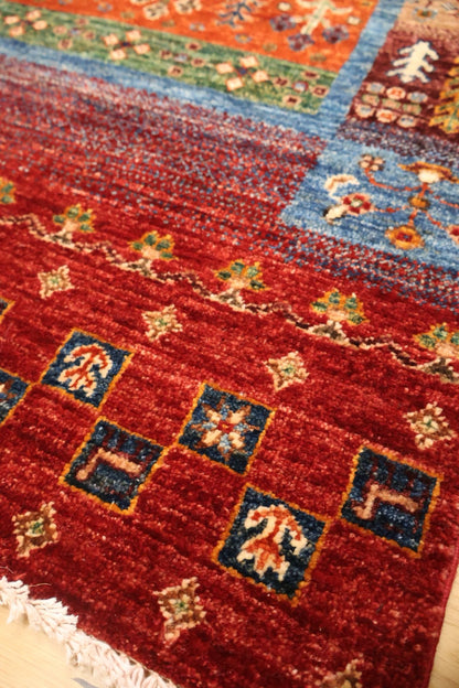 A close-up of a patterned area rug with a deep red field, blue borders, and small beige and green floral motifs.