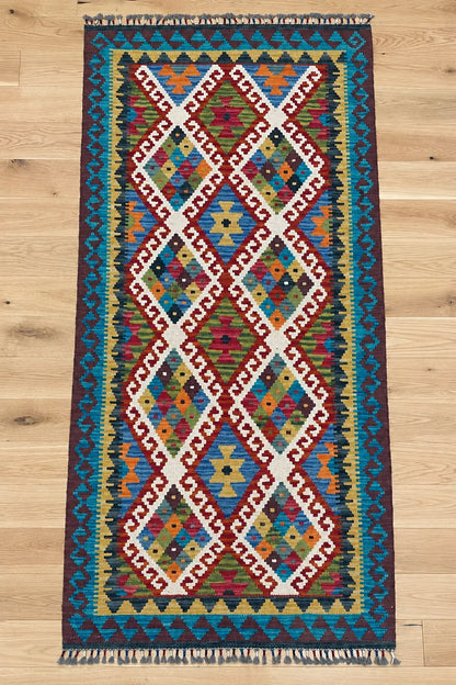 A long rectangular kilim runner rug with a vibrant geometric diamond pattern, displayed flat on a light wood floor.