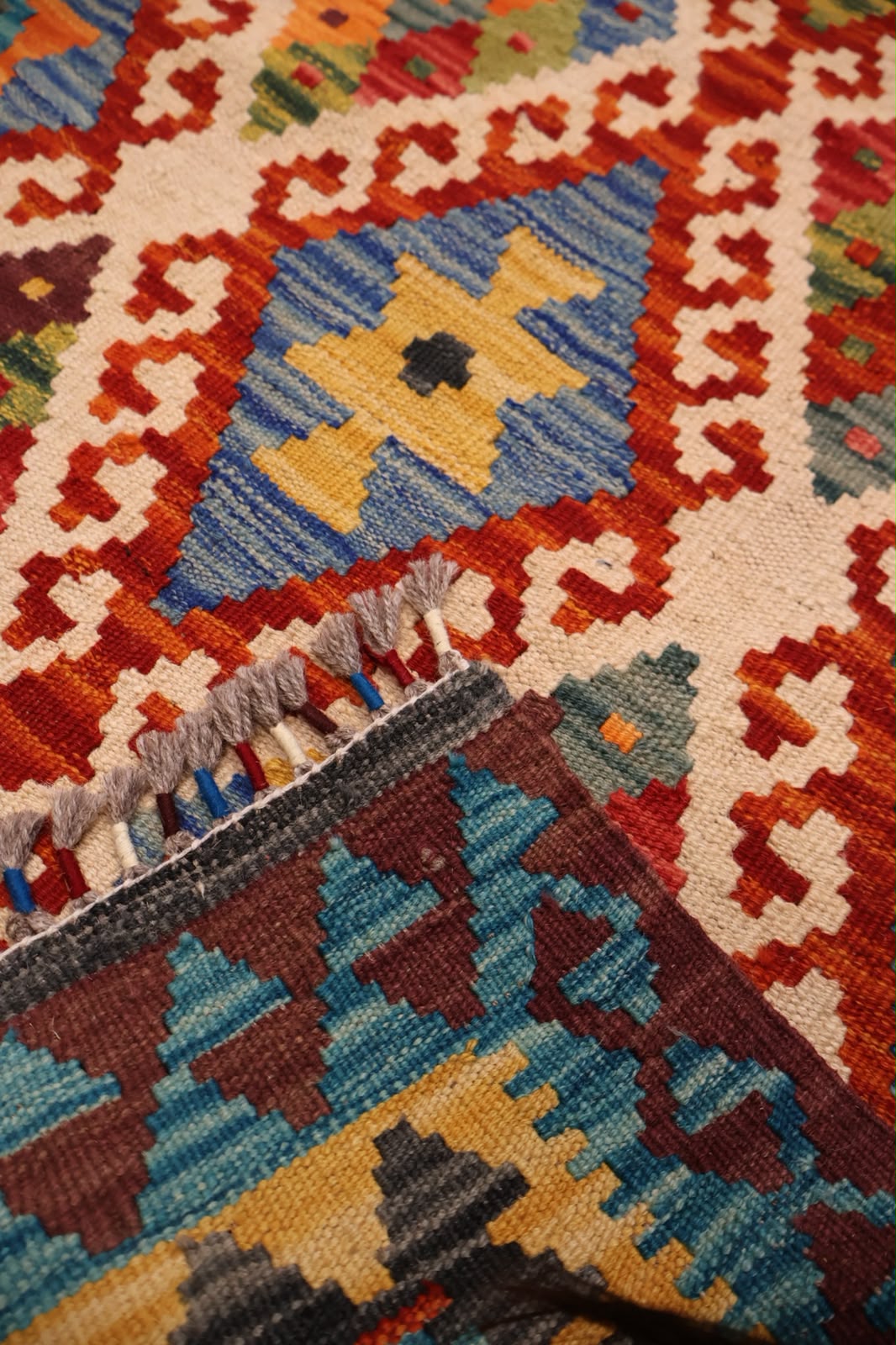 Close-up of a woven geometric rug with a cream base, red and orange border patterns, a central blue diamond with a yellow cross motif, and multicolored fringe.
