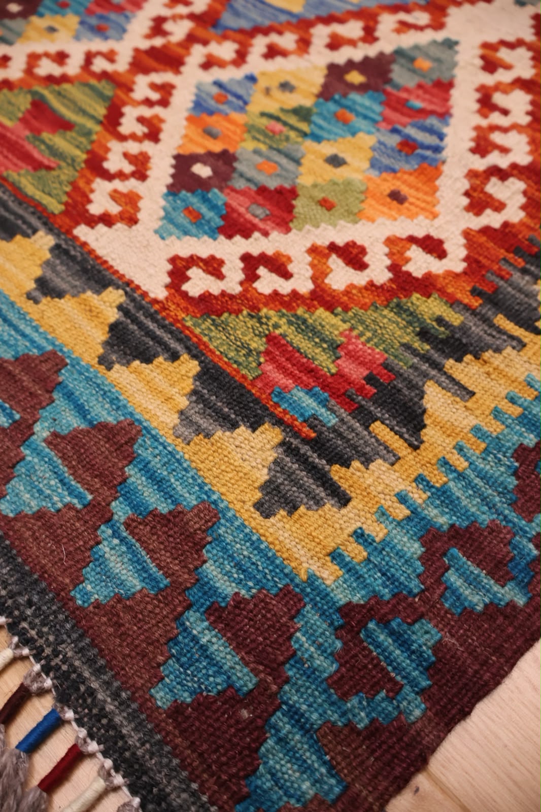 A close-up of a colorful woven rug with geometric patterns in blue, red, yellow, brown, black, and cream, showing the texture and fringe detail.
