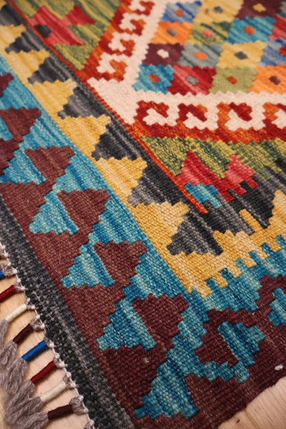 Close-up of a colorful geometric kilim rug with a woven pattern and fringed edge, showing shades of blue, red, yellow, brown, black, and cream.