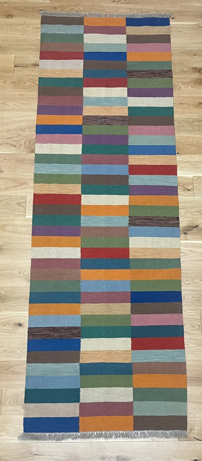 A long rectangular woven rug with a multicolor patchwork stripe pattern, laid flat on a light wood floor.