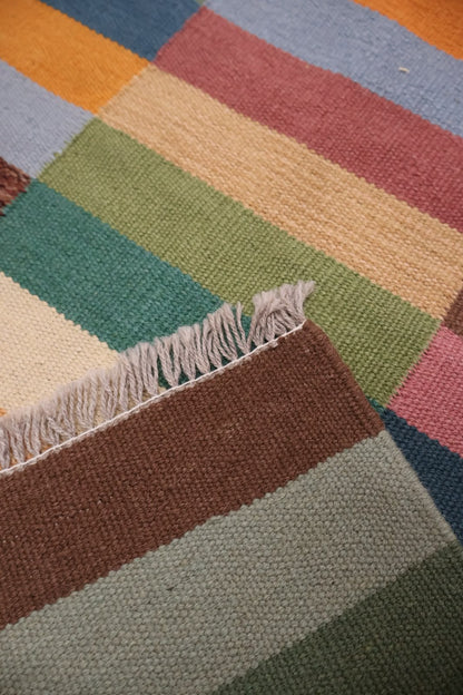 Close-up of a woven rug with a striped patchwork design in earthy tones including brown, sage green, beige, teal, pink, and blue, featuring a fringed edge.