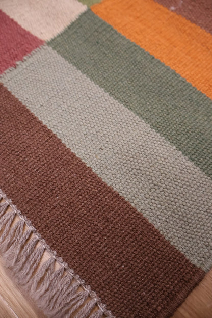 A close-up of a woven rug with broad horizontal stripes in muted brown, beige, sage green, orange, and dusty pink, finished with a fringed edge.