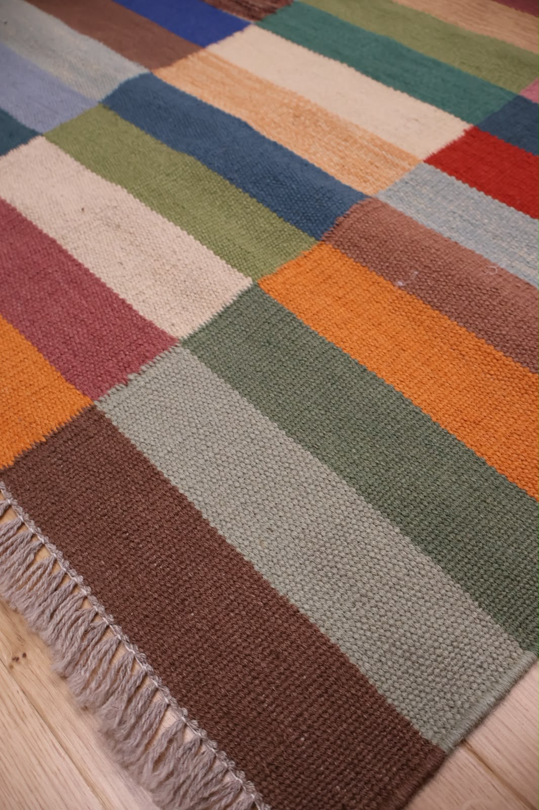 A close-up of a woven patchwork rug with multicolored rectangular blocks and a fringed edge, laid on a light wood floor.