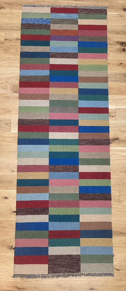 A long rectangular woven rug with a multicolor patchwork stripe pattern, laid flat on a light wood floor.