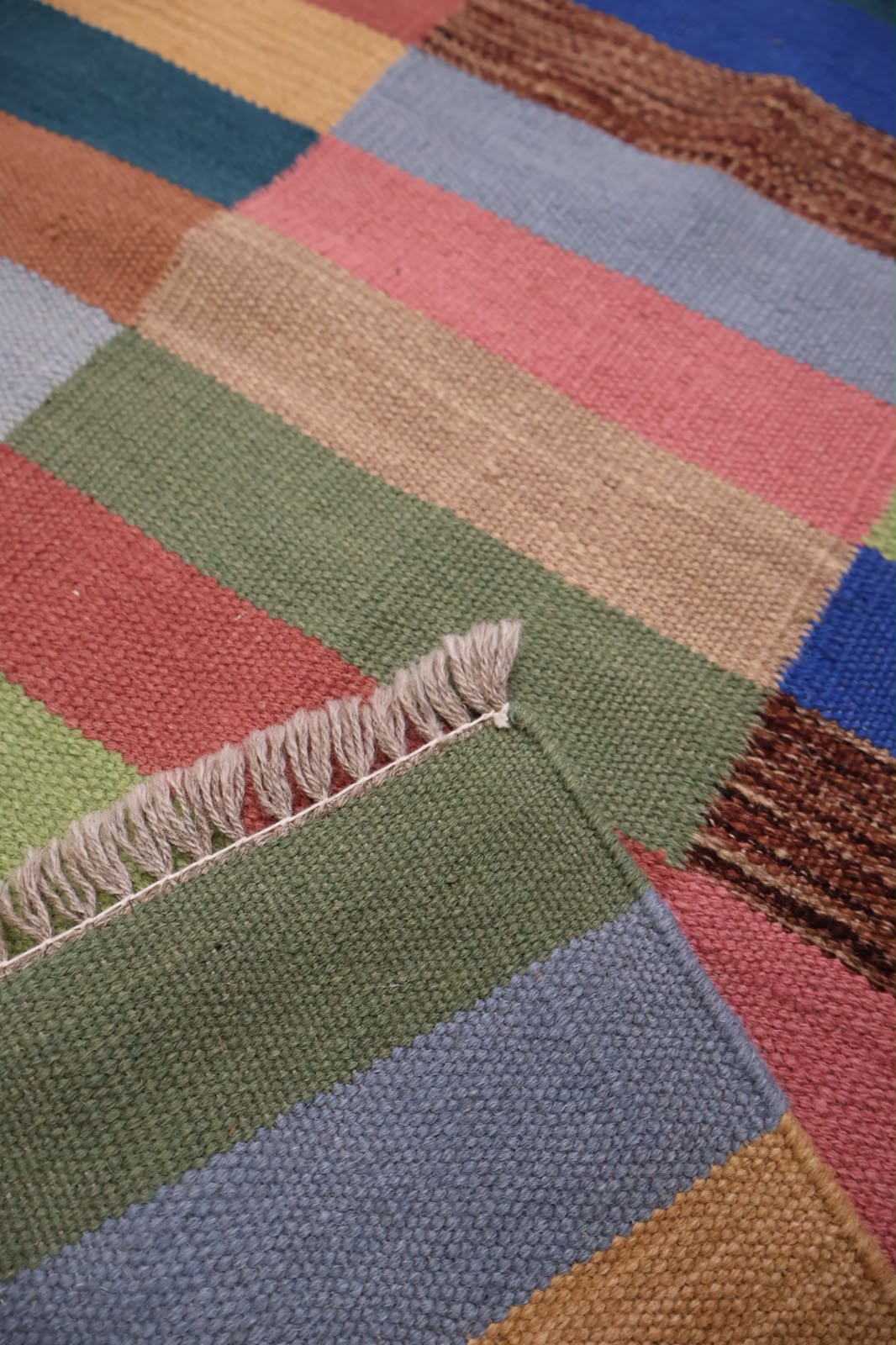 Close-up of a woven textile with broad multicolored stripes in muted green, dusty blue, pink, tan, mustard, and brown, finished with short fringe on the edge.