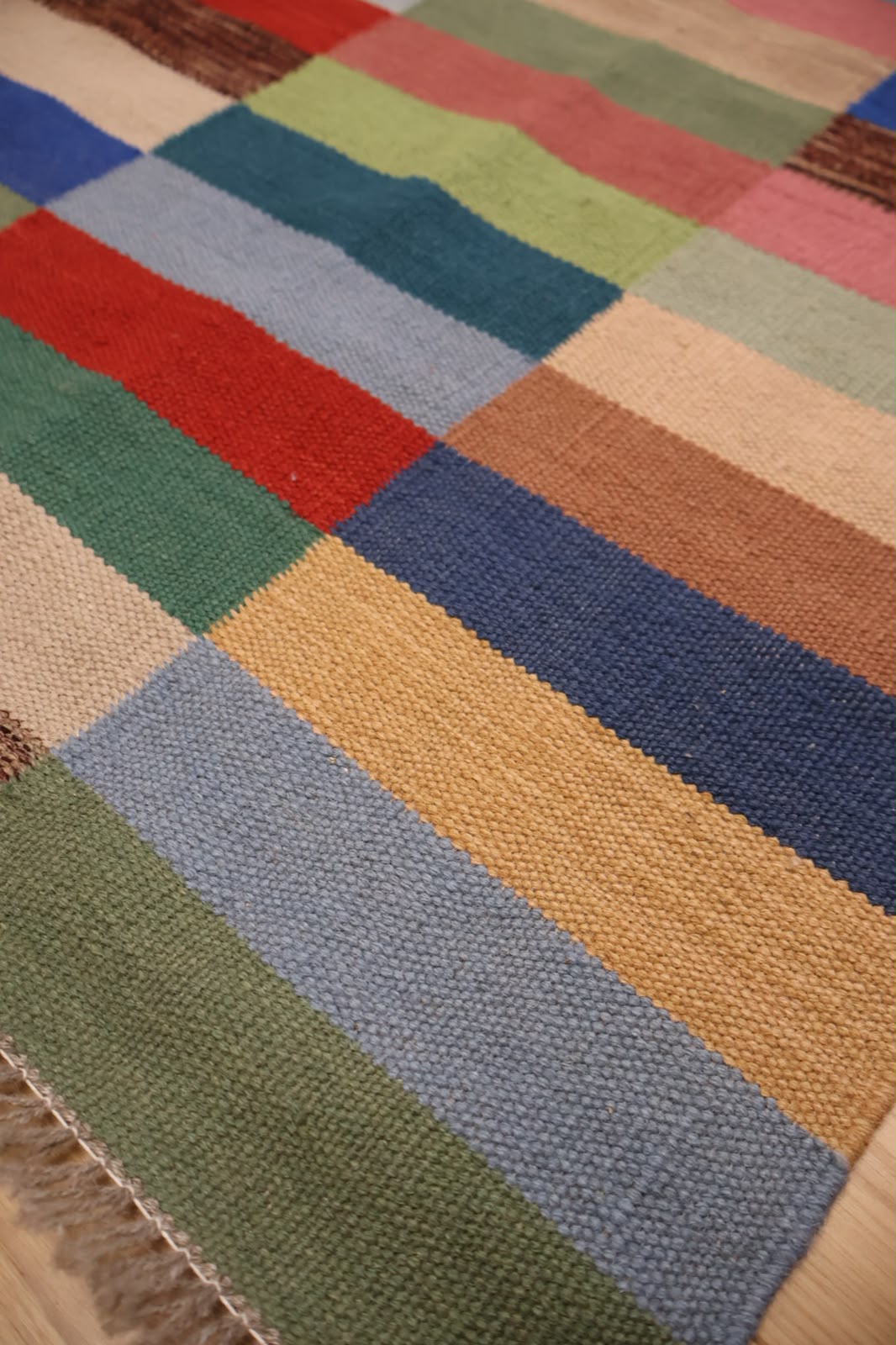 A close-up of a woven patchwork rug with rectangular blocks of green, blue, red, beige, yellow, and pink, showing a textured natural fiber weave and fringe along one edge.