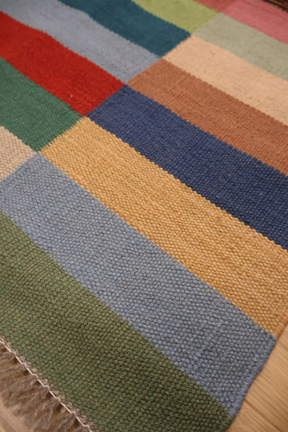 A close-up of a woven rug with a patchwork design of colored stripes, including green, red, blue, beige, and mustard yellow, with a fringed edge visible.