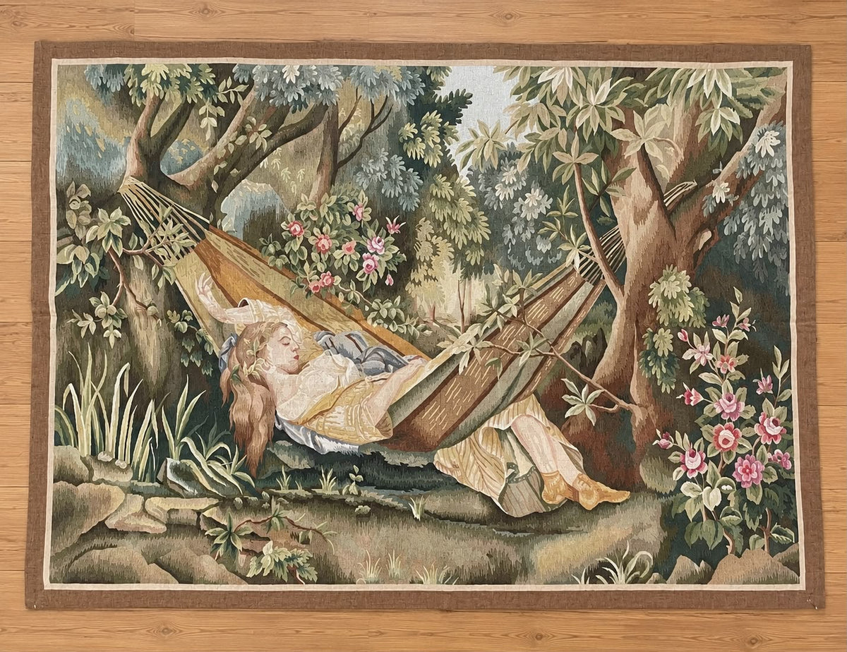 A rectangular tapestry featuring a vintage-style illustration of a woman relaxing in a hammock surrounded by lush green trees and pink flowers, displayed flat on a light wood floor.