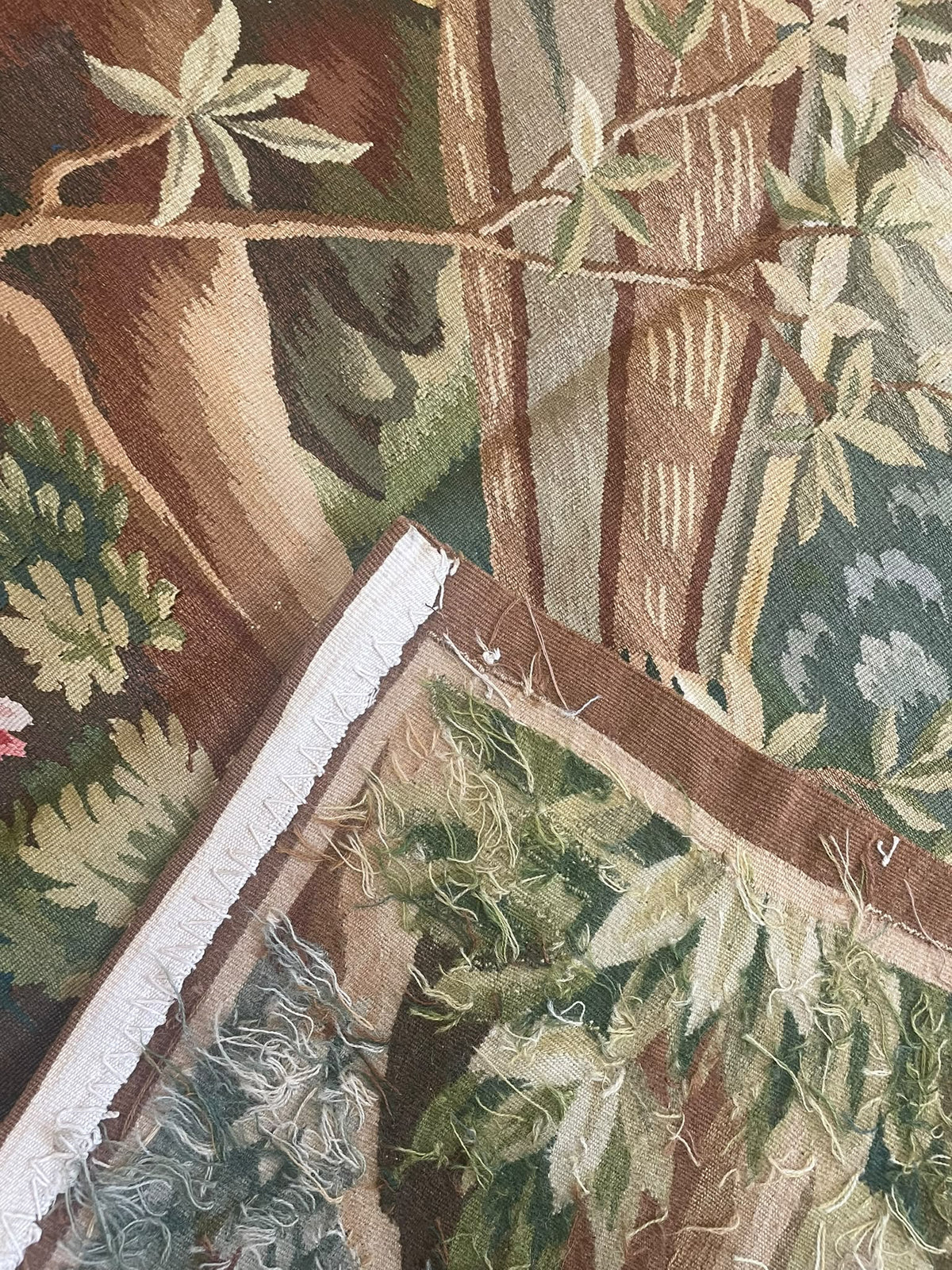 A close-up of a woven tapestry featuring a botanical and tree pattern in earthy tones of brown, green, beige, and cream, with visible fringe along one edge.
