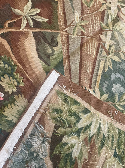 A close-up of a woven tapestry featuring a botanical and tree pattern in earthy tones of brown, green, beige, and cream, with visible fringe along one edge.