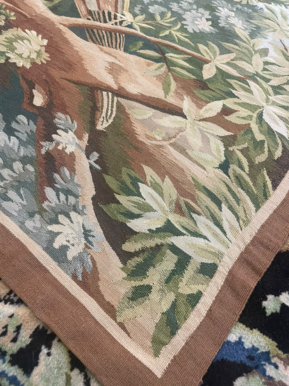 A close-up of a woven tapestry showing a botanical and tree motif with green leaves, brown branches, cream flowers, and a tan border, laid over a dark patterned textile.