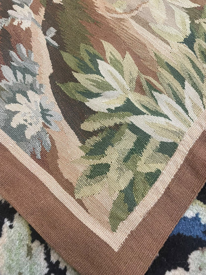 Close-up of a woven tapestry corner showing a botanical pattern with green leaves, cream flowers, and a brown border.