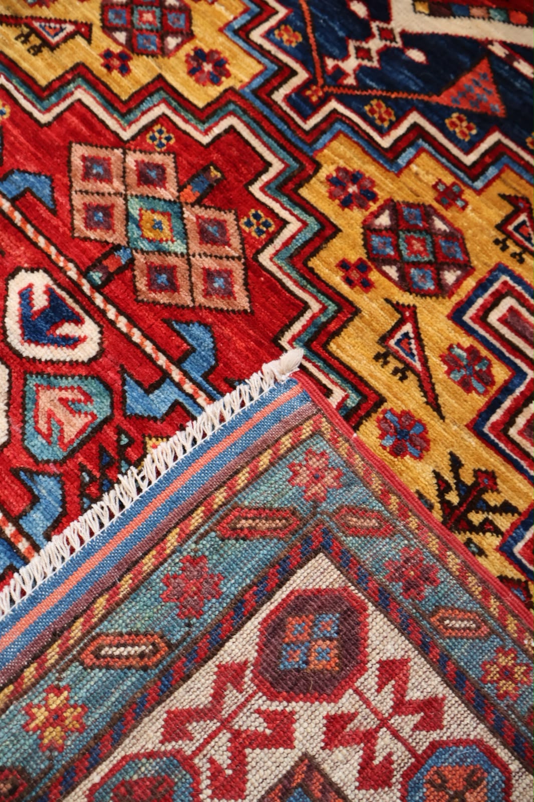 Close-up of a patterned area rug showing a corner with white fringe, featuring geometric motifs in red, yellow, blue, cream, and brown.