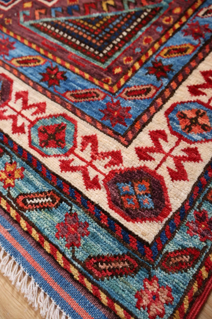 A close-up of a traditional patterned rug showing its corner detail, featuring cream, blue, red, and black geometric motifs with white fringe.