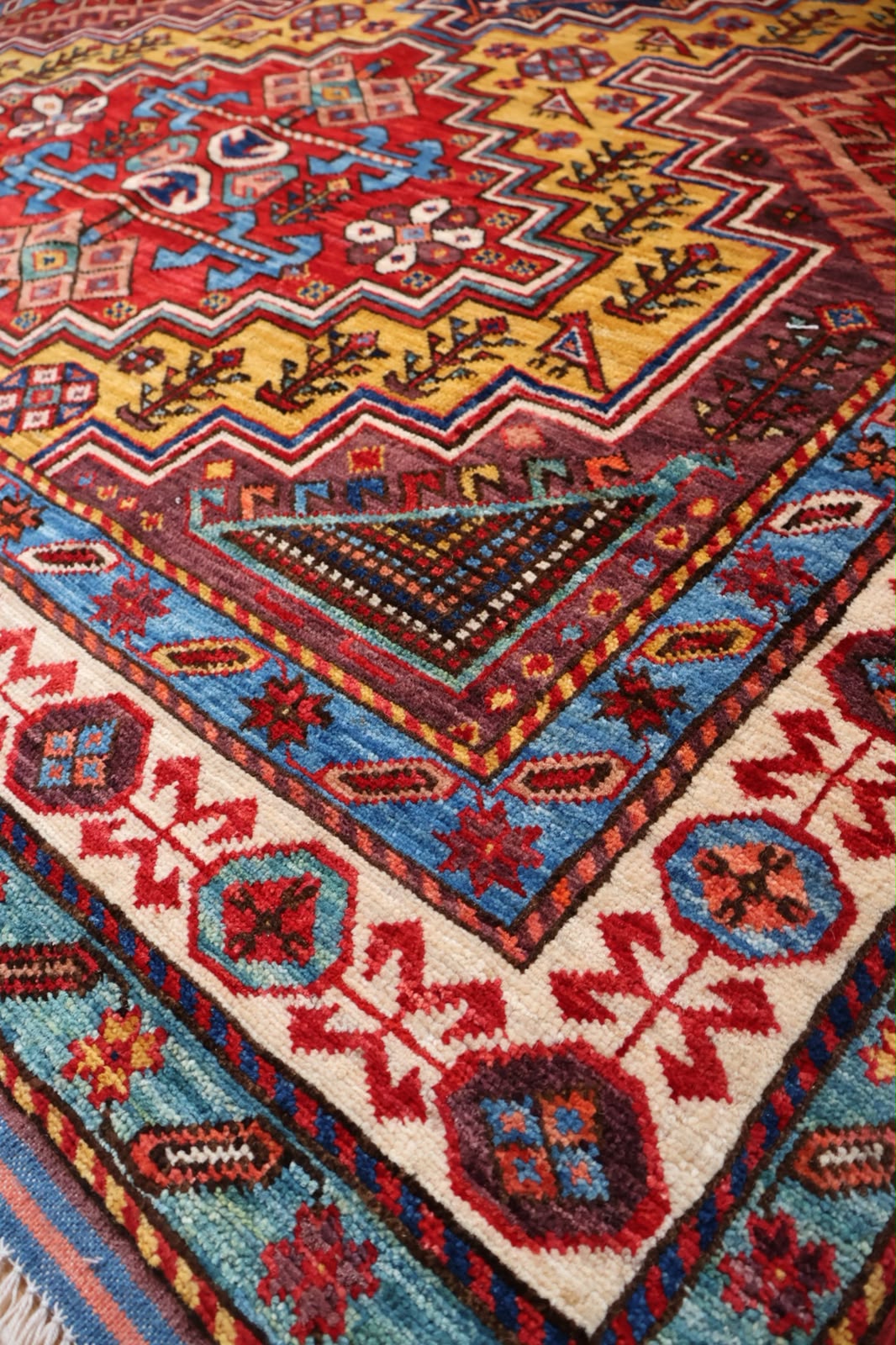 A close-up of a traditional patterned rug showing detailed woven geometric motifs in red, blue, yellow, beige, and brown.
